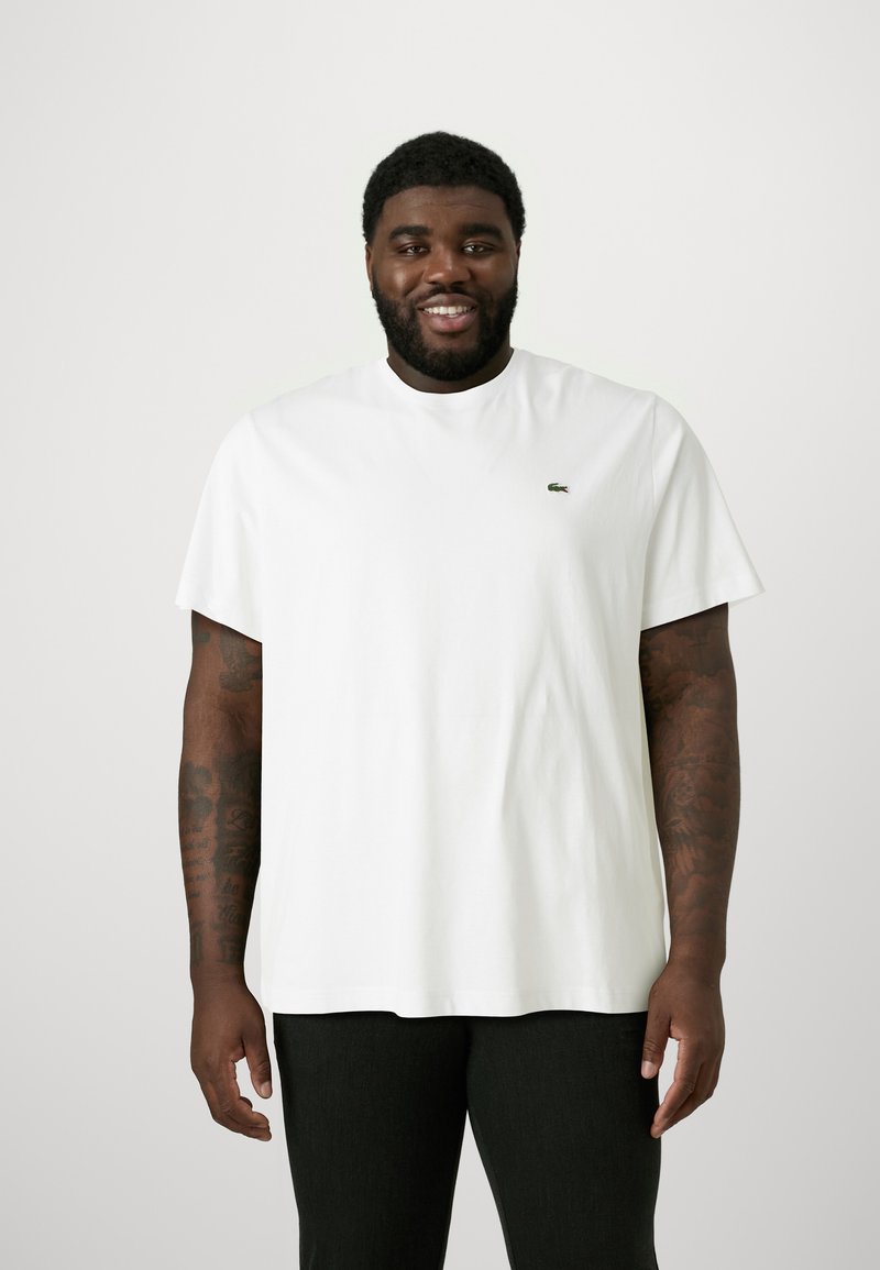 Camiseta de algodón blanca con cuello redondo y mangas cortas; presenta un pequeño logo verde de Lacoste en el lado izquierdo del pecho.