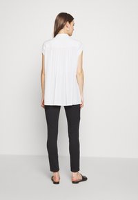 Blusa branca de mangas curtas com colarinho, apresentando um corte solto e pregas nas costas. Combinada com calças pretas justas e sandálias pretas.