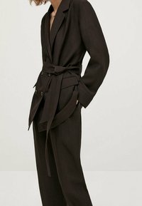 Blazer marron foncé, cintré à la taille, à double boutonnage et avec de larges revers ; porté avec un pantalon assorti.