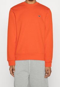 Persona che indossa una felpa a maniche lunghe di colore arancione brillante con un piccolo logo di coccodrillo bianco e nero sul petto e pantaloni grigi chiari.