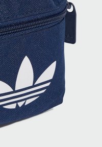 Tmavomodrá taška vyrobená z textúrovanej látky, s veľkým bielym geometrickým logom a tromi horizontálnymi pruhmi blízko spodnej časti. Zipsové zapínanie.