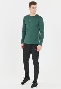 Sportliches, langärmliges grünes Oberteil mit strukturiertem Muster und schwarzen Sporthosen mit Cargo-Taschen und einem Logo, kombiniert mit schwarzen Turnschuhen.