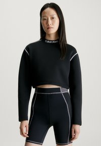 Maglione corto nero con collo alto e cuciture argentate, abbinato a pantaloni ciclistici neri con dettagli grigi e una vestibilità strutturata.