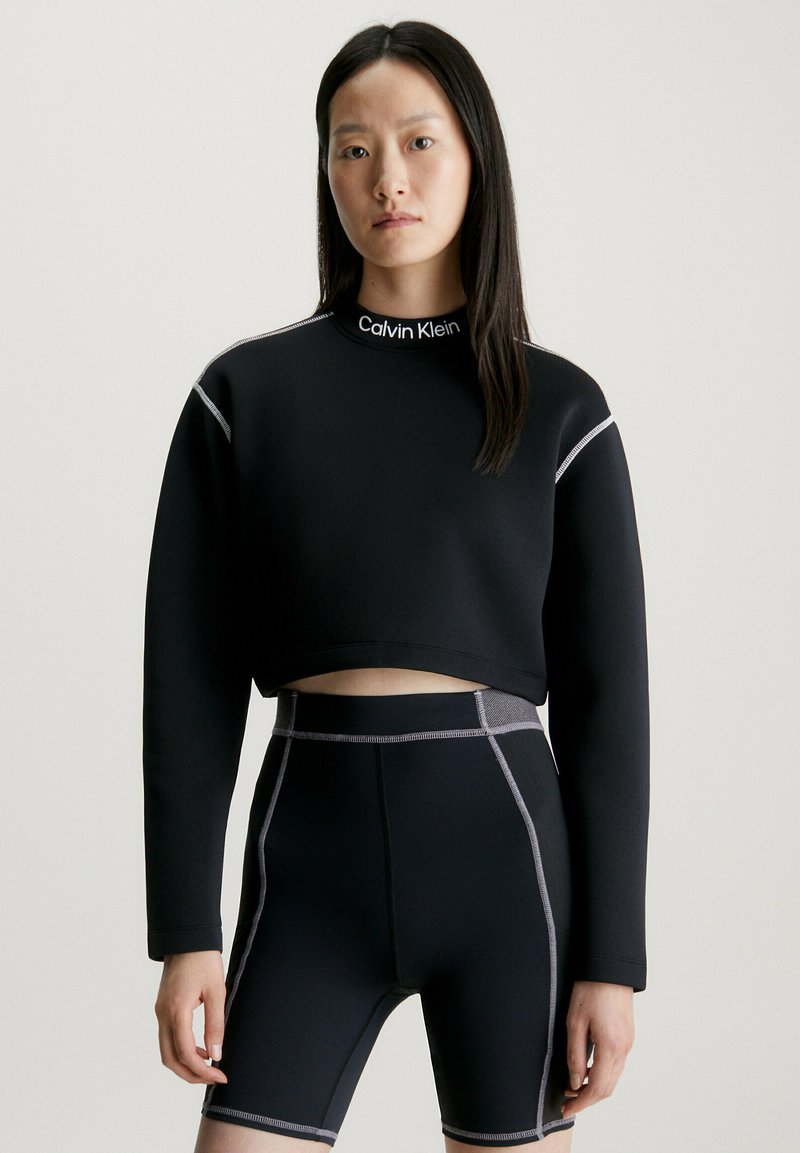 Maglione corto nero con collo alto e cuciture argentate, abbinato a pantaloni ciclistici neri con dettagli grigi e una vestibilità strutturata.