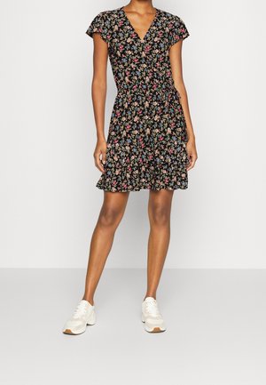 Kvinde iført sort blomsterprintet slå-om-kjole med korte ærmer og flæset kant, kombineret med hvide sneakers, stående foran en ensfarvet baggrund.