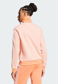 Ružičasti dukserica s visokim ovratnikom i dugim rukavima, s glatkom površinom. Donji rub odgovara hlačama s bijelim prugama.