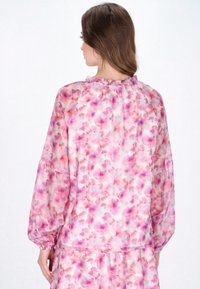 Blusa fluida in tessuto leggero e trasparente con un motivo floreale rosa e viola. Presenta un colletto arruffato e maniche a palloncino.
