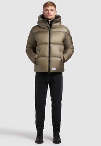 Puffer jacket in olive green, featuring a hood, quilted design, front zipper, and side pockets. Paired with black jeans and boots.