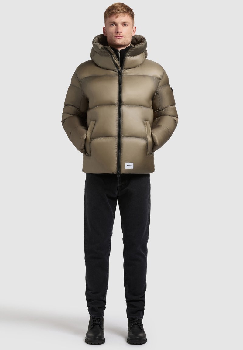 Puffer jacket in olive green, featuring a hood, quilted design, front zipper, and side pockets. Paired with black jeans and boots.