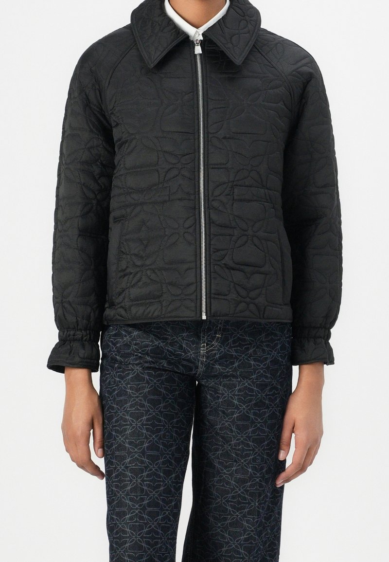 Personne portant une veste matelassée noire avec une fermeture éclair, associée à un pantalon bleu foncé à motifs, debout devant un fond blanc uni.
