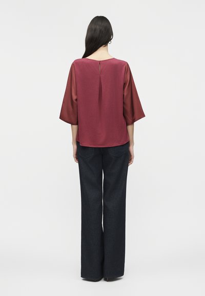 Γυναίκα με μακριά σκούρα μαλλιά, που φοράει μια φαρδιά μπορντό μπλούζα με διαφανή μανίκια τριών τετάρτων και σκούρο παντελόνι με φαρδιά μπατζάκια, όρθια με την πλάτη γυρισμένη.