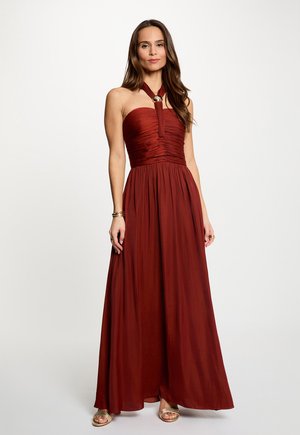 Donna con un abito lungo fino al pavimento, color bordeaux, senza spalline, con dettaglio halter e sandali dorati, in piedi davanti a uno sfondo chiaro uniforme.