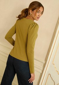 Zielona oliwkowa bluzka z długim rękawem i bufkami na ramionach. Gładka tekstura, dopasowany krój i okrągły dekolt. Połączona z ciemnym dżinsem.