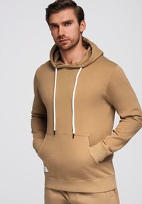 Homme avec une barbe châtain clair portant un sweat à capuche beige avec des cordons blancs, mains dans la poche avant, sur un fond clair uni.