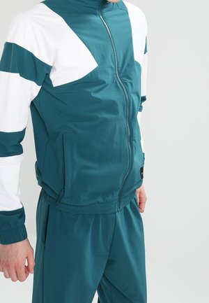 Homme portant une veste de sport zippée turquoise et blanche et un pantalon turquoise assorti, debout avec la main gauche détendue le long du corps.