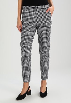 Femme portant un pantalon slim noir et blanc à motif pied-de-poule avec des talons slingback noirs, debout avec une main dans la poche.
