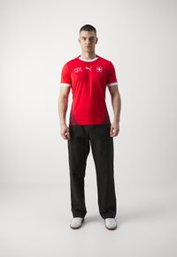 Rød atletisk trøje med hvide accenter, prydet med et grafisk motiv på brystet og et schweizisk flag. Kombineret med sorte bukser og hvide fodtøj.