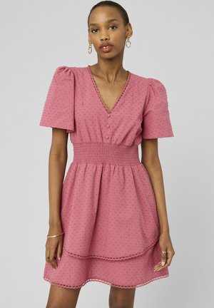 Femme portant une robe rose texturée à manches courtes avec des boutons, une taille froncée et un ourlet festonné, debout devant un fond uni.