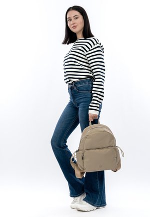 Beige rygsæk med to lynlåse foran og topstrop, båret af en person i en stribet skjorte og blå jeans, stående foran en hvid baggrund.