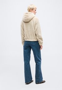 Άτομο με κοντά ξανθά μαλλιά, που φοράει μπεζ χνουδωτό φούτερ με κουκούλα, μπλε τζιν και καφέ παπούτσια, στέκεται και κοιτάζει μακριά προς μια απλή φόντο.