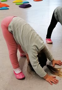 Ein Kind in einem grauen Sweatshirt und pinken Leggings beugt sich nach vorne und berührt den Boden. In der Nähe sind bunte geometrische Formen auf einer grauen Fläche verstreut.