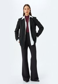 Zwarte blazer met pieklapellen, witte blouse eronder, bordeauxrode coltrui en zwarte flared broek. Glad materiaal, minimale details.