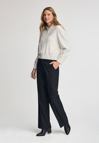 Hellgrauer Sweatshirt mit einem Viertel-Reißverschluss-Kragen, geripptem Saum und besticktem Logo. Kombiniert mit schwarzen weiten Hosen und Ankle Boots.