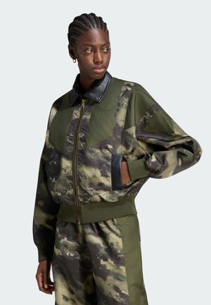 Persona che indossa una giacca mimetica verde e nera con zip e colletto in pelle e pantaloni abbinati, guarda di lato con la mano in tasca.