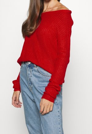Femme portant un pull en maille rouge ample décolleté aux épaules et un jean taille haute bleu clair, debout devant un fond uni.