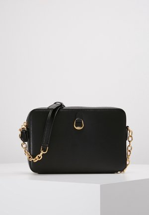 Sac bandoulière rectangulaire en cuir noir avec sangle chaîne dorée et petit détail anneau doré à l'avant, présenté sur surface blanche.