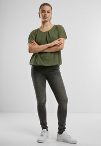 Olivgrüne Bluse mit kurzen Ärmeln und rundem Halsausschnitt sowie Falten, kombiniert mit dunkelgrauen Skinny Jeans und weißen Sneakern.