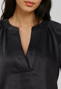 Nahaufnahme einer Frau, die eine schwarze Satinbluse mit gerafftem Einsatz und V-förmigem Ausschnitt trägt, die Schultern und oberen Brustbereich zeigt.