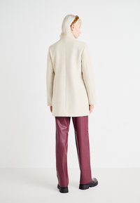 Beige ullblandad kappa med rakt snitt, bakslits och utan synliga knäppningar, i kombination med burgundy imitation läderbyxor.