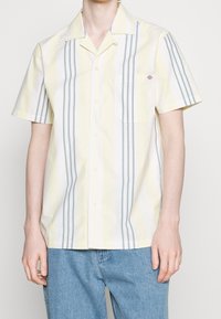 Uomo che indossa una camicia a maniche corte, con bottoni, a righe verticali gialle e grigie e una tasca sul petto sinistro, abbinata a jeans blu.