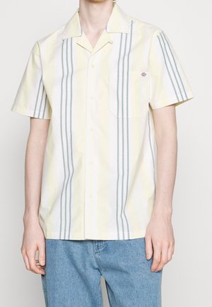Man in een kortogige, knoopsluiting shirt met verticale gele en grijze strepen en een zak op de linkerborst, in combinatie met blauwe jeans.