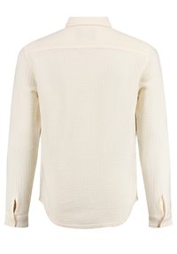 Camisa de manga larga color beige claro con una tela texturizada, cuello estándar, puños abotonados y aberturas laterales. Vista trasera.
