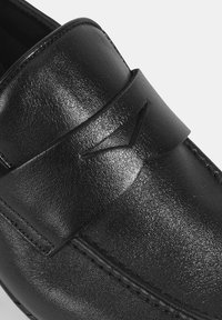 Zwarte leren instapper met een glanzende afwerking, gestikte details en een decoratieve band met een subtiele logo-detail.