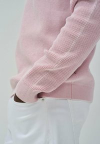 Maglione rosa chiaro con texture, orlo e polsini a costine, abbinato a jeans bianchi con un accento di bottone argentato sulla tasca.