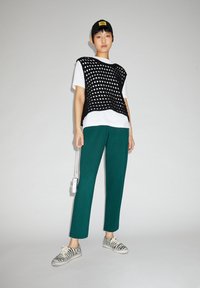 Zwarte gebreide vest met witte stippen over een wit T-shirt, gecombineerd met groene broek en schoenen met zebra-strepen. Zwarte pet met geel logo.