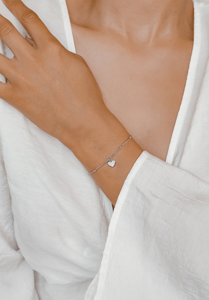 Pulsera de cadena de plata con un pequeño colgante en forma de corazón, llevada en una mano. El fondo presenta una suave tela blanca, resaltando el diseño de la pulsera.