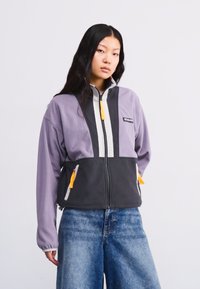Fleece jacket in purple and black, featuring a zip front, high collar, and contrasting yellow accents. Front pockets with zippers.