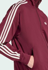 Bordový zipový kabát s vysokým golierom, biele trojpruhové akcenty na rukávoch a bočné vrecká. Hladká textúra a atletický strih.