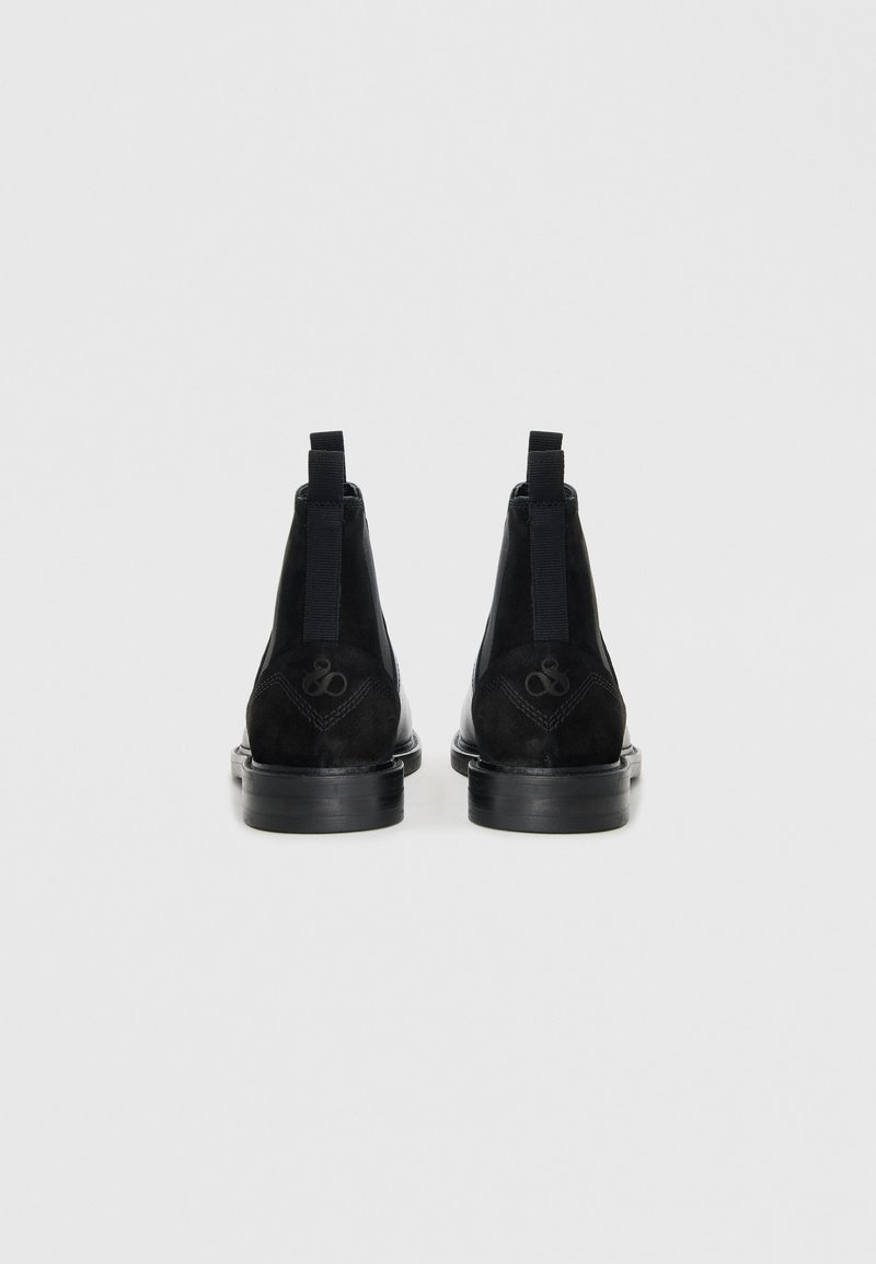 Schwarze Wildleder-Ankle-Boots mit elastischen Seiteneinsätzen, verstärktem Absatz und flacher Gummisohle. Auffälliges geprägtes Logo auf der Rückseite.