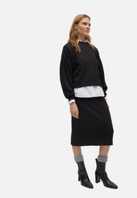 Sudadera corta negra superpuesta sobre una camisa blanca, combinada con una falda lápiz negra. La modelo lleva botas de tacón negras con calcetas arrugadas grises.