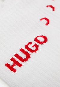 Fehér szövet, amelyen vörös hímzett szöveg olvasható: "HUGO." A textúra puhanak tűnik, és enyhén bordázott mintázat van az felületen.