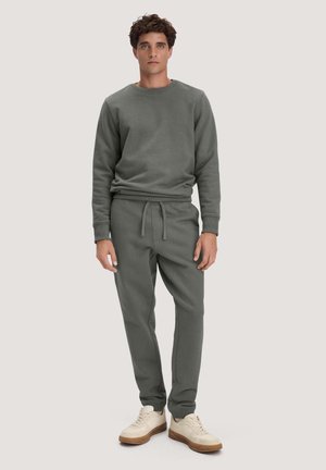 Man staat met een donkergrijze trui en bijpassende joggingbroek met beige sneakers op een effen lichte achtergrond.