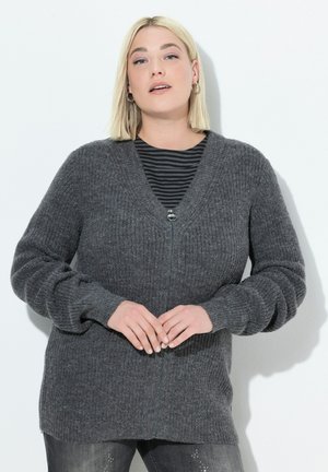 Nainen, jolla on lyhyet vaaleat hiukset, käyttää tummanharmaata vetoketjullista neulepuseroa mustan ja harmaan raidallisen paidan päällä, seisoo valkoista taustaa vasten.