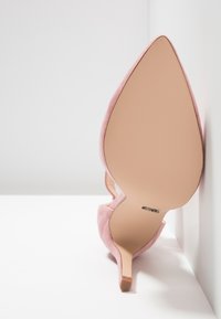 Chaussure à talon haut en daim rose à bout pointu vue de la semelle, avec un talon stiletto fin reposant contre une surface blanche.