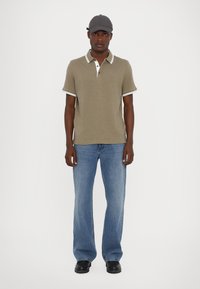 Kortermet olivengrønn polo-shirt med hvite detaljer, kombinert med lyseblå vide jeans og svarte sko. Modell med grå caps.