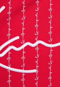 Röd tyg med vita broderade mönster, inklusive snirkliga linjer och slingor, som skapar en texturerad och mönstrad yta.